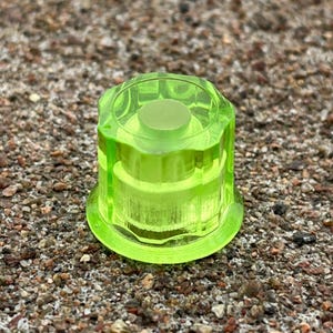 Puede incluir: Un pomo de color amarillo neón translúcido con un diseño geométrico de múltiples facetas, una parte superior circular y una base plana más ancha. El objeto está fabricado con un material transparente similar al vidrio que deja pasar la luz.