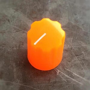May include: A bright orange cylindrical control knob featuring a scalloped, ridged edge for grip and a single thin white indicator line printed on the top surface. The knob has a smooth, matte finish and a circular base, designed for electronic equipment.