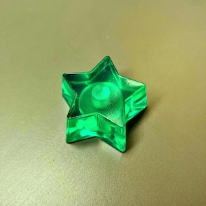 May include: A transparent, green star-shaped object with a circular design in the center. The star has five points and is made of a glossy material. The object is sitting on a neutral-toned surface.
