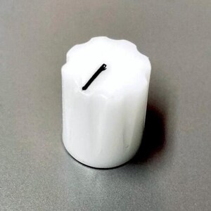 May include: A cylindrical white knob with a fluted, scalloped edge design. The top surface features a single, thin, black diagonal line marking. The knob has a smooth, glossy finish and a vertical orientation, showing a consistent white color throughout.