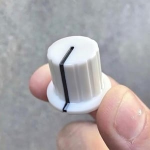 May include: A white control knob with a black line indicator. The knob has a cylindrical shape with a ridged edge and a flat base. The knob is held in a person's hand.