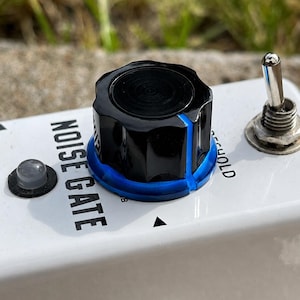 May include: A white guitar effects pedal featuring a black control knob with a blue base, a silver toggle switch, and a small circular indicator light. The text NOISE GATE is printed in black on the white surface next to the knob.