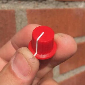 Könnte beinhalten: Ein roter Bedienknopf mit einer weißen Markierungslinie. Der Knopf ist zylindrisch mit einem leicht breiteren Sockel. Das Bild ist eine Nahaufnahme, die die Details des Designs und der Farbe des Knopfes hervorhebt.