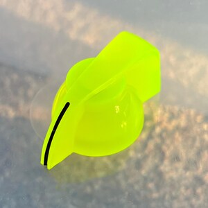 May include: A neon yellow plastic control knob with a circular base and a pointed indicator arm. The indicator arm features a thin, vertical black line running along its side to mark the position of the dial on electronic equipment.