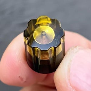 May include: A small, cylindrical knob with a translucent, dark amber-colored top featuring a scalloped, gear-like edge. The base of the knob is a solid, opaque dark brown color, and the center of the top displays a bright, reflective yellow circular detail.