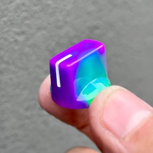 Pode incluir: Um botão roxo e verde com uma linha branca na frente. O botão é feito de plástico e tem um acabamento liso e brilhante.