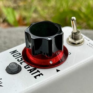 Pode incluir: Um pedal de efeitos de guitarra branco com a etiqueta NOISE GATE, apresentando um grande botão de controlo preto sobre uma base vermelha metálica, uma pequena luz indicadora cinzenta, um interruptor de metal prateado e uma entrada jack.