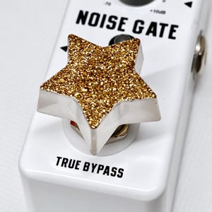 May include: A white guitar effects pedal labelled NOISE GATE and TRUE BYPASS, featuring a prominent star-shaped control knob with a textured gold glitter finish on its top surface, mounted on a white metal chassis with black printed text.