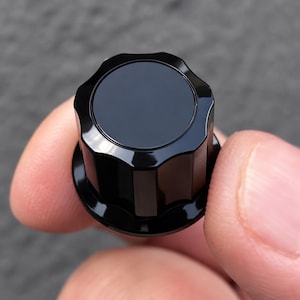 May include: A glossy black control knob featuring a circular top surface and a fluted, scalloped side profile with vertical ridges. The knob has a smooth, reflective finish and a wide base, designed for adjustment on electronic equipment.
