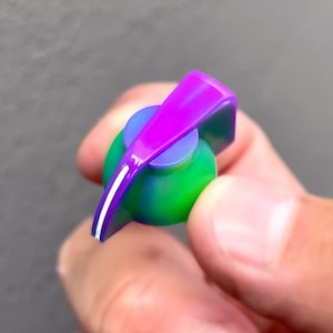 Op de afbeelding: Een paarse en groene knop met een witte lijn in het midden. De knop is gemaakt van plastic en heeft een gladde, glanzende afwerking.