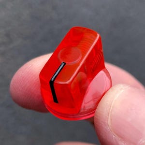 May include: A translucent red plastic control knob featuring a cylindrical base and a flat top surface. A single vertical black indicator line is printed on the side and top edge of the knob to show the current setting or position clearly.