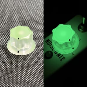 Puede incluir: Un botón verde translúcido con una parte superior en forma de estrella, diseñado para un dispositivo electrónico. El botón brilla intensamente en la oscuridad, con el texto "NOISE GATE" visible en el dispositivo.