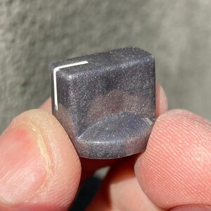 May include: A dark gray control knob with a glittery, shimmering finish and a single white indicator line on the top edge. The knob has a D-shaped profile with a flat front face and a curved back, designed for adjusting electronic equipment settings.