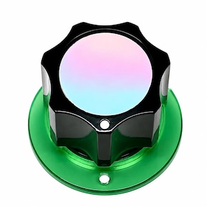 Puede incluir: Un pomo negro en forma de estrella con un centro liso e iridiscente que cambia entre tonos rosas y azules. El pomo está montado sobre una base circular de color verde translúcido que presenta un pequeño orificio centrado en el borde inferior.