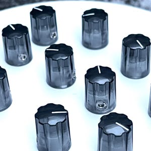 May include: A collection of black, cylindrical control knobs with a ridged design and a white indicator line on top. Each knob has a small hole on the side. The knobs are arranged on a white surface.