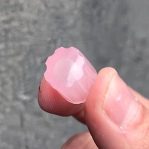 Op de afbeelding: Een gefacetteerde, lichtroze edelsteen die tussen vingers wordt gehouden. De edelsteen heeft een unieke, bloemachtige vorm met meerdere zijden. De kleur is een zacht, doorschijnend roze en de randen zijn duidelijk gedefinieerd.