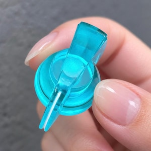 May include: A translucent bright turquoise plastic pump dispenser head featuring a circular base and a downward-angled spout. The component is designed for liquid dispensing, showcasing a smooth, glossy finish and a clear, vibrant cyan color throughout the entire structure.