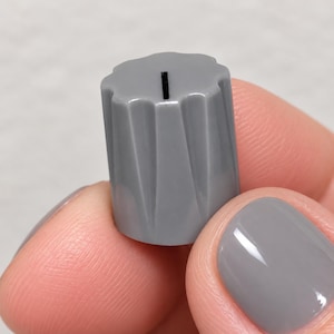 Peut inclure: Un bouton de commande cylindrique gris doté d'un design extérieur cannelé et festonné avec des crêtes verticales. La surface supérieure du bouton est plate et comprend une fine ligne indicatrice verticale noire centrée pour indiquer la position.