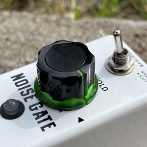 May include: A white guitar effects pedal labeled NOISE GATE with a black and translucent green control knob, a silver metal toggle switch, and a small circular indicator light. The text THRESHOLD is printed on the white surface near the knob.