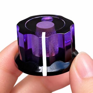 May include: A translucent purple plastic control knob with a scalloped edge and a solid black base. The top surface features a circular indentation, and a single vertical white line is printed on the side for precise adjustment.