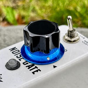 May include: A white guitar effects pedal featuring a black knob with a blue metallic base. The text NOISE GATE is printed in black on the top surface. A silver toggle switch, a small circular indicator light, and a metal input jack are visible.