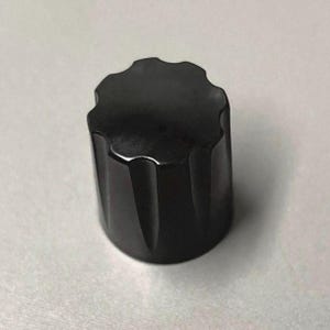 Può includere: Una manopola di controllo cilindrica nera solida caratterizzata da un bordo superiore smerlato simile a un ingranaggio con scanalature verticali lungo i lati. La manopola presenta una finitura opaca liscia e una superficie superiore circolare.
