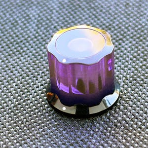 May include: A close-up of a purple and silver knob. The knob has a circular top with a white centre and a silver rim. The body of the knob is a translucent purple with vertical ridges. The base is a shiny silver colour.
