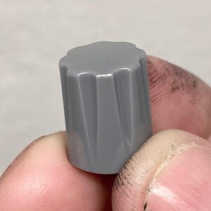 May include: A cylindrical gray plastic control knob featuring a series of vertical fluted grooves along its exterior surface. The top edge of the knob has a scalloped design, providing a textured grip for manual adjustment of electronic equipment.