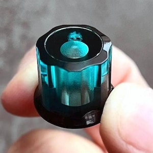 Könnte beinhalten: Ein zylindrischer Knopf aus durchscheinendem blaugrünem Material mit gewelltem Oberrand. Der Knopf hat eine glatte, glänzende Oberfläche, eine vertiefte Mitte und einen dunklen, massiven Sockel, wodurch die innere Struktur sichtbar wird.