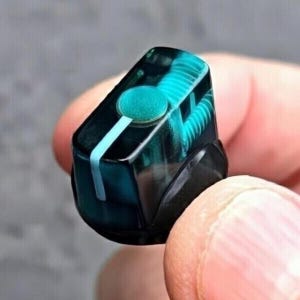 May include: A translucent dark teal knob with a cylindrical shape and a flat top surface. The knob features a bright cyan circular center and a single vertical cyan line extending from the center to the bottom edge, encased in clear resin.
