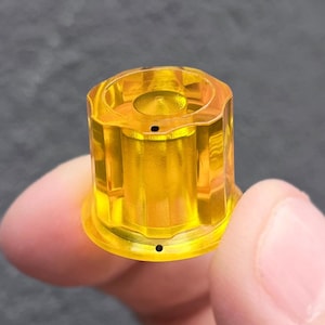 May include: A translucent yellow cylindrical knob featuring a fluted exterior design with vertical ridges. The top surface has a recessed center and a small black dot indicator, while the base includes a matching black dot on the outer rim.