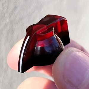 May include: A translucent dark red guitar control knob with a tapered pointer shape. The knob features a smooth, glossy finish and a distinct white indicator line running along the side of the pointer, designed for precise adjustment of audio levels.