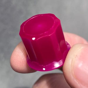 May include: A small, cylindrical, magenta-colored plastic cap featuring a fluted or ribbed exterior design with a smooth, flat top surface and a slightly wider, circular base rim, showing a glossy finish with subtle light reflections.