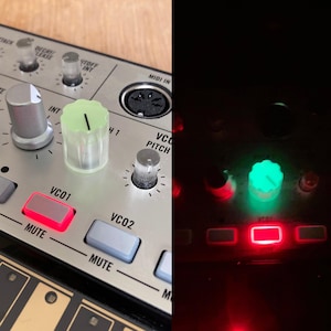 May include: A synthesizer control panel featuring a silver metallic surface with various knobs and buttons. One prominent knob is translucent pale green and glows bright green in low light. A rectangular button labeled VC01 glows red.