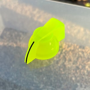 Op de afbeelding: Een felgele plastic knop met een zwarte lijn in het midden. De knop heeft de vorm van een afgeronde pijlpunt.