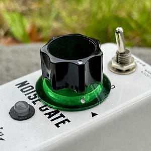 May include: A white guitar effects pedal labeled NOISE GATE with a large black knob featuring a green translucent base. The pedal includes a silver metal toggle switch, a small circular grey indicator light, and a metallic input jack port.