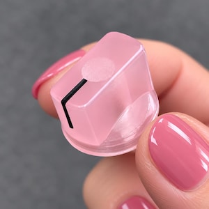 May include: A translucent light pink knob with a smooth, rounded rectangular top and a circular indentation. A single thin black vertical line extends from the center of the top edge down the front face of the knob, indicating position.