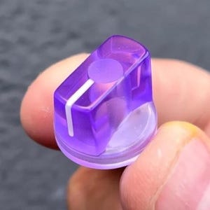 May include: A translucent purple plastic control knob with a circular top and a flat side. The knob features a single white vertical indicator line running from the center top down the side, designed for precise adjustment on electronic equipment.