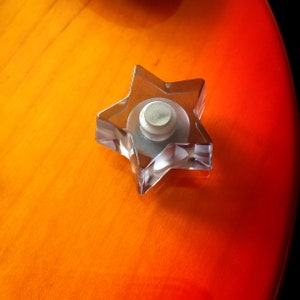 Op de afbeelding: Een doorzichtige stervormige knop met een ronde, zilveren knop in het midden. De knop bevindt zich op een feloranje oppervlak.