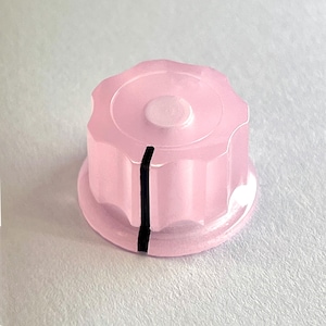 Peut inclure: Un bouton en plastique rose avec une ligne noire au centre. Le bouton a un bord festonné.