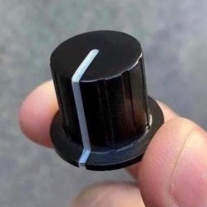 Puede incluir: Un mando de control cilíndrico de color negro con una línea indicadora blanca vertical que va desde el borde superior hasta la base. La superficie exterior del mando tiene una serie de estrías verticales para el agarre y una base circular.