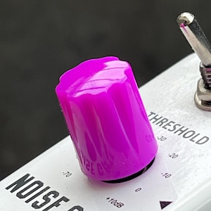 Könnte beinhalten: Nahaufnahme eines violetten Knopfes an einem weißen Noise-Gate-Pedal. Der Knopf ist mit "Threshold" beschriftet und hat Markierungen von -30 bis 0. Der Text "Noise Gate" ist unterhalb des Knopfes sichtbar.