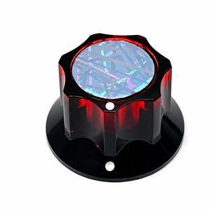 Puede incluir: Un pomo rojo translúcido con forma de estrella de seis puntas y una base circular. La superficie superior presenta un inserto holográfico con un patrón geométrico de líneas que se cruzan y reflejan la luz en varios colores.