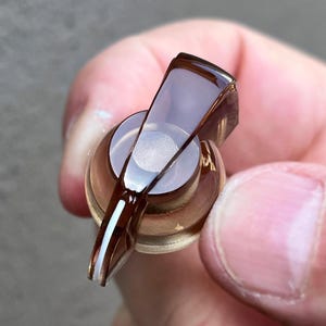 May include: A translucent dark brown control knob with a smooth, glossy finish and a tapered pointer. The knob features a circular base and a raised, elongated top section with a white line indicator running along the edge of the pointer.