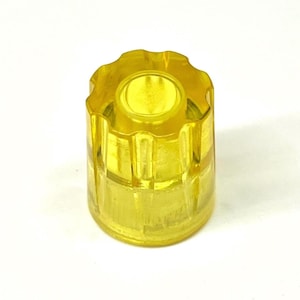 May include: A translucent yellow cylindrical control knob featuring a scalloped, gear-like top edge and a smooth, straight-sided base. The material is clear, allowing light to pass through the structure, revealing a hollow center and internal details.