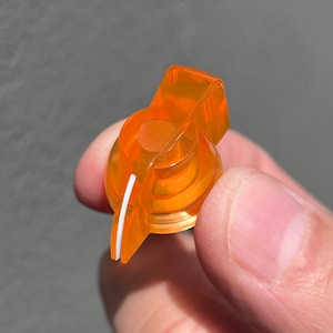 Op de afbeelding: Oranje plastic knop met een witte lijn in het midden. De knop is ontworpen voor gebruik met een gitaar of ander muziekinstrument.