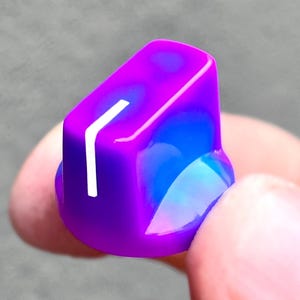 May include: A glossy control knob featuring a vibrant gradient of purple and blue. The knob has a smooth, rounded cylindrical shape with a flat top surface and a single, vertical white indicator line printed on the side for precise adjustment.