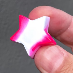 Może przedstawiać: Gwiezdny zawieszek z żywicy w odcieniach różu i bieli. Gwiazda jest trójwymiarowa i ma gładkie, błyszczące wykończenie.