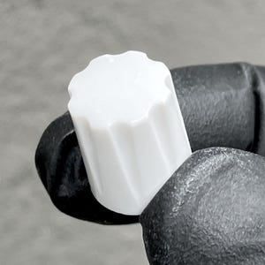 May include: A small, cylindrical white plastic control knob featuring a fluted or ribbed exterior design with vertical grooves. The knob has a flat top surface with a scalloped edge, providing a textured grip for manual adjustment of equipment.