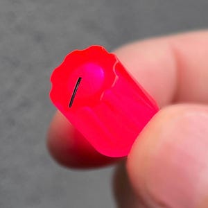 May include: A bright neon pink control knob with a scalloped edge and a vertical black indicator line on the top surface. The knob features a cylindrical body with vertical ridges along the sides, designed for adjustment on electronic equipment.